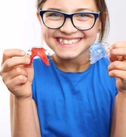Girl wearing glasses holding red and blue retainers and smiling.