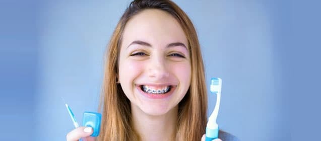 Young woman with braces smiling while holding toothbrushes for orthodontic care.