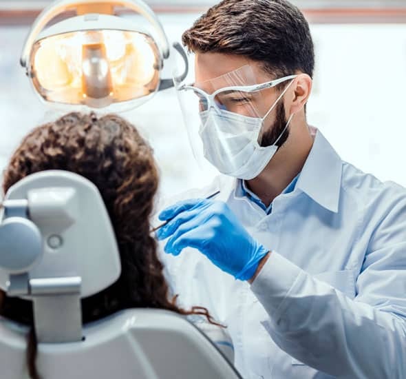 Dentist in white coat and protective gear examining patient in dental chair.