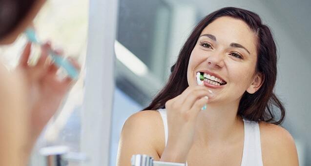 Woman brushing her teeth and smiling at mirror during morning dental routine.