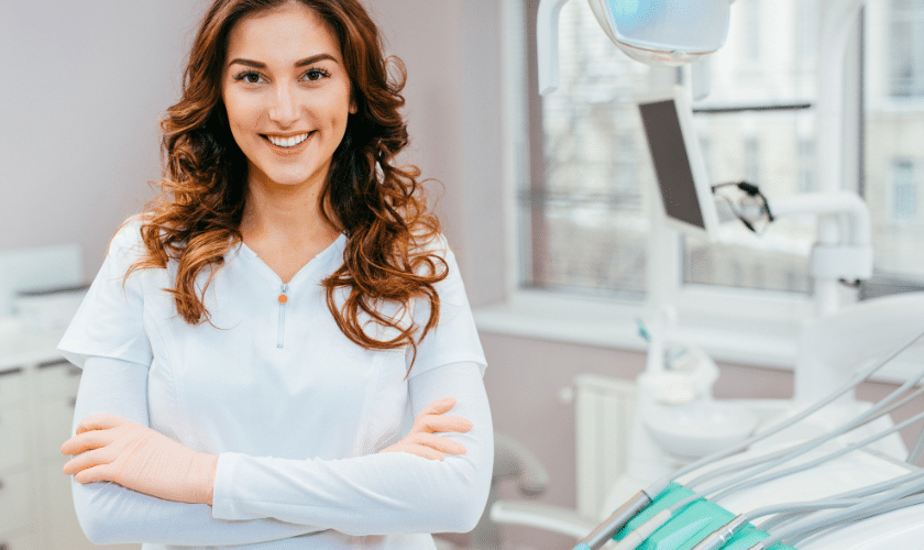 Smiling female dental professional in white coat standing in modern dental office treatment room.