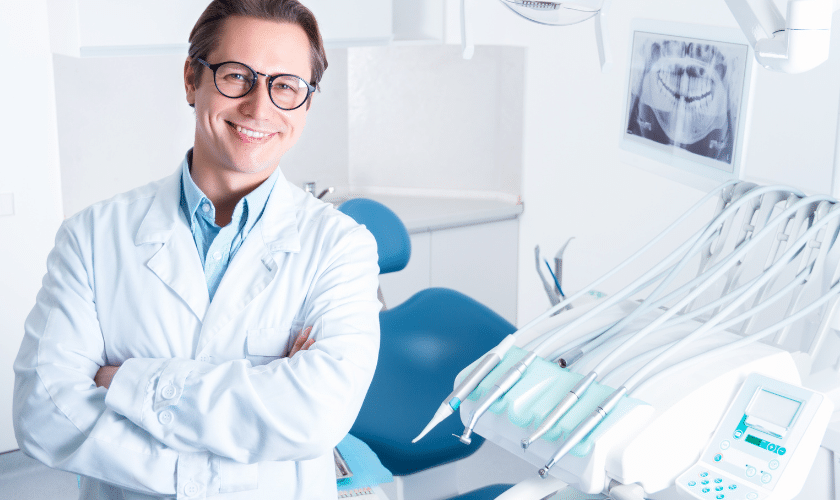 Smiling dentist in white coat standing in modern dental office with treatment chair and X-rays.