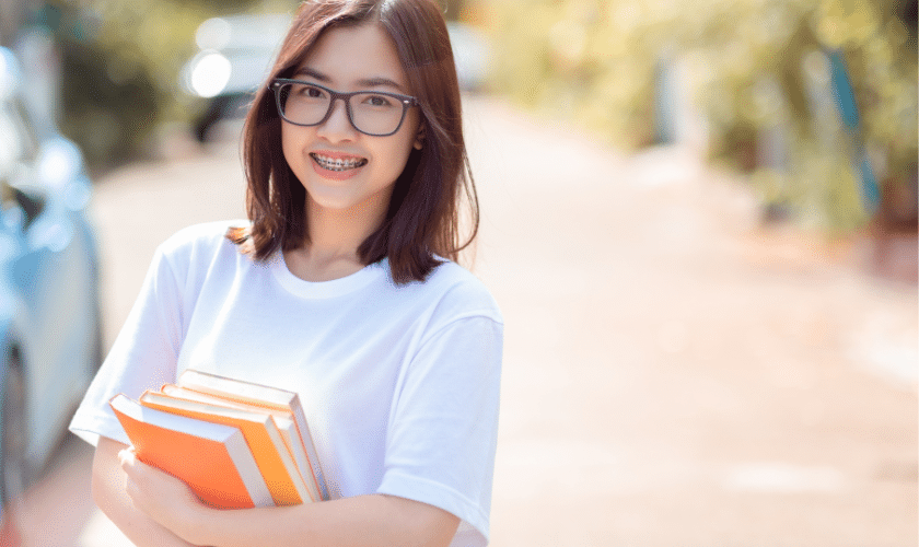 Young student wearing glasses holding textbooks, smiling outdoors.