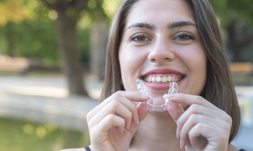 Smiling woman holding clear Invisalign aligners to show teeth straightening treatment.