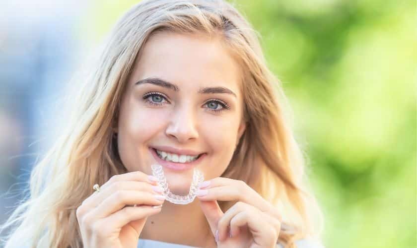 Young woman smiling while holding clear orthodontic aligners for teeth straightening treatment.