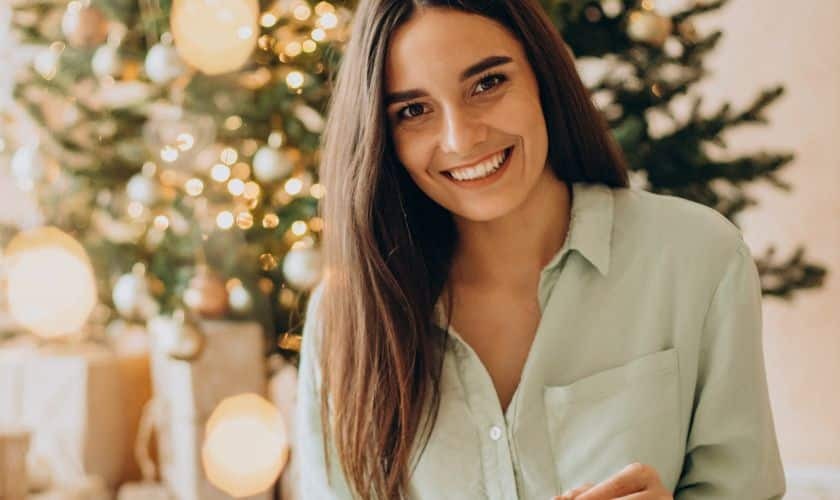 Woman with radiant smile near Christmas tree, representing holiday dental care with Invisalign.
