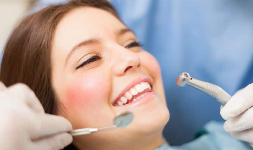 Woman smiling during pediatric dental examination with dentist holding instruments.