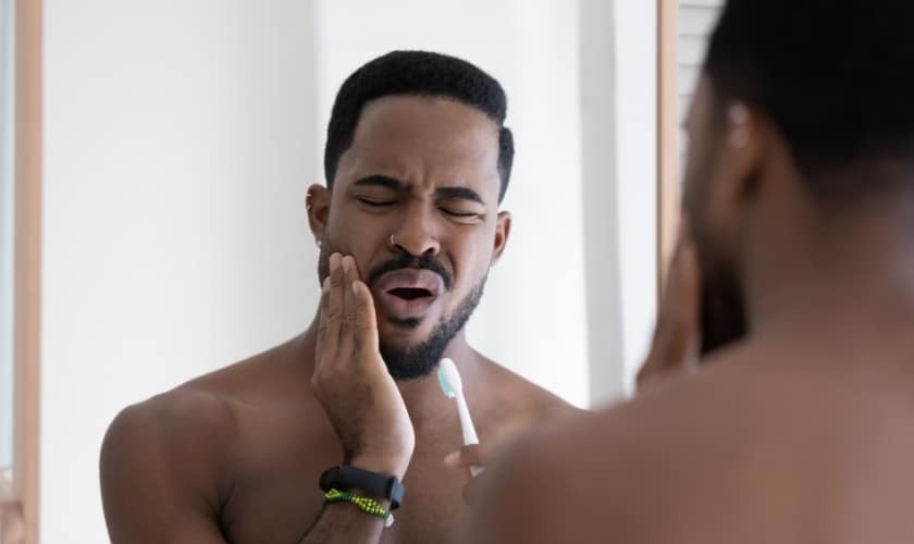 Man experiencing tooth pain while brushing, touching his cheek near his jaw.