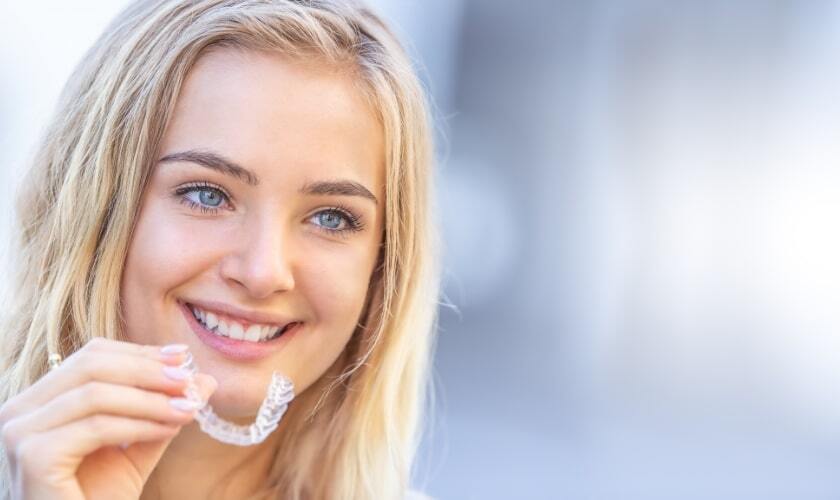 Young woman holding clear aligner tray and smiling at camera for Invisalign treatment.