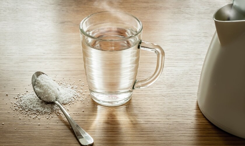 Saltwater gargle remedy setup with glass of water, spoon with salt, and pitcher on wooden surface.