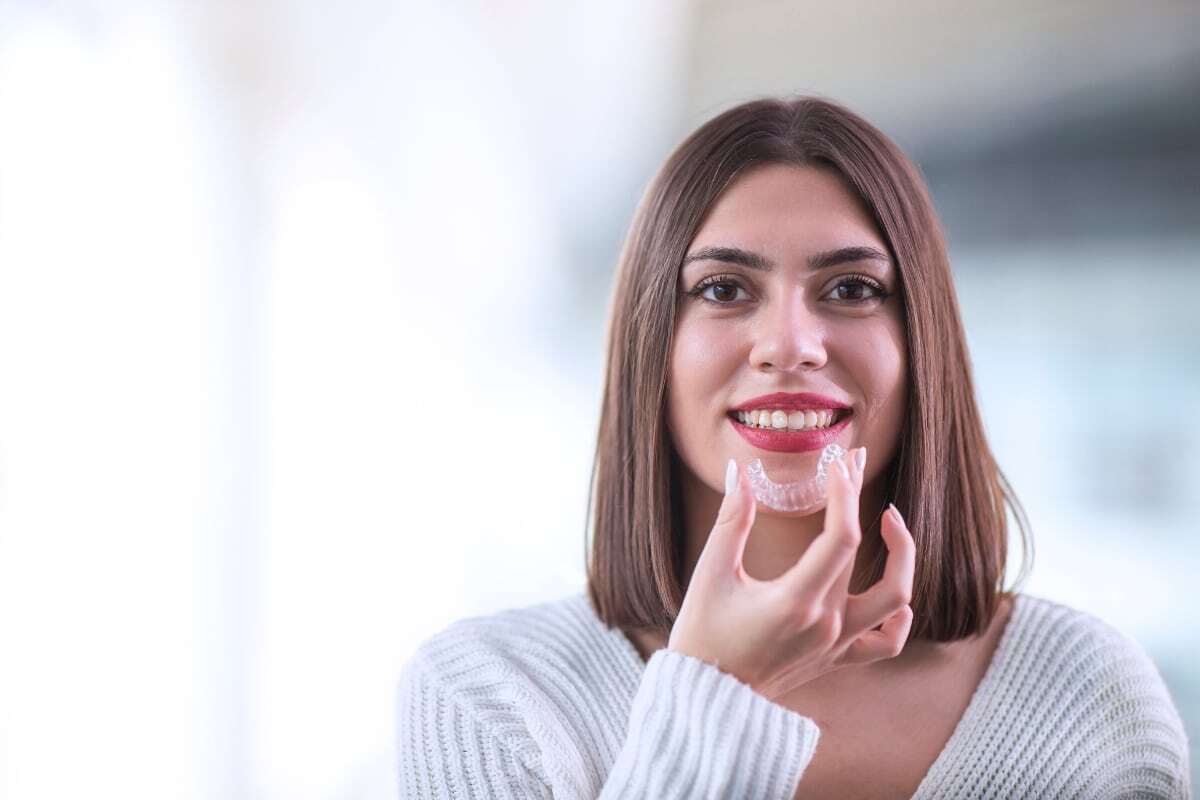 Woman holding clear Invisalign aligner and smiling to show teeth correction results.