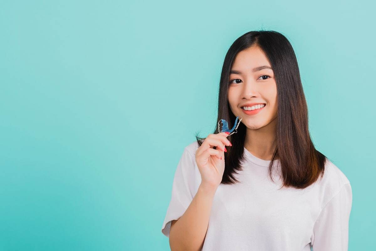 Young woman holding orthodontic retainer while smiling against turquoise background.