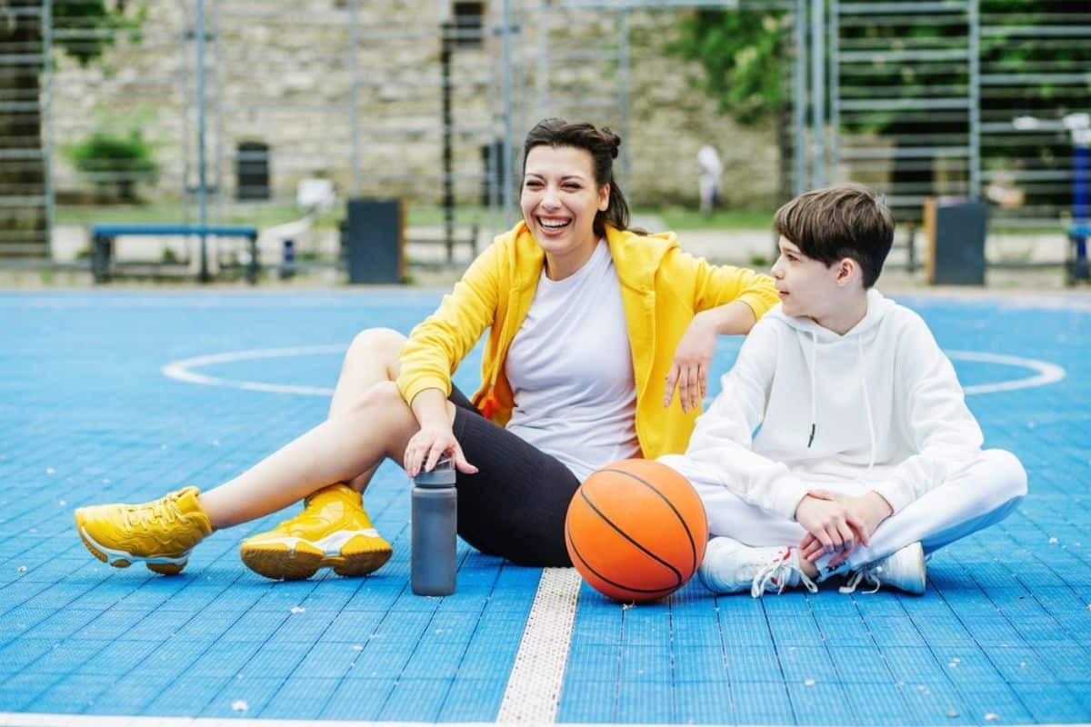 Woman and boy sitting on basketball court with basketball and water bottle, demonstrating active sports participation.