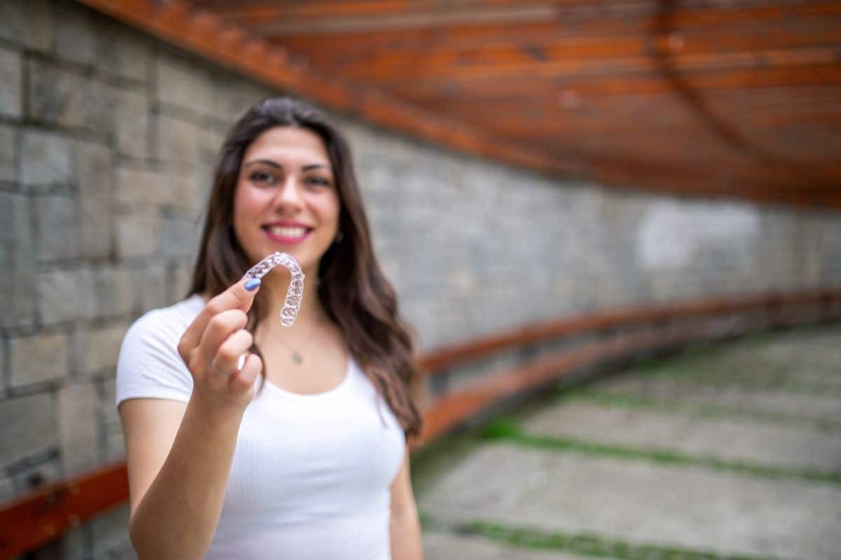 Young woman holding clear Invisalign aligner and smiling outdoors.