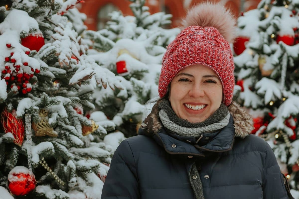Woman in red knit hat smiling outdoors with snow-covered Christmas trees behind her.