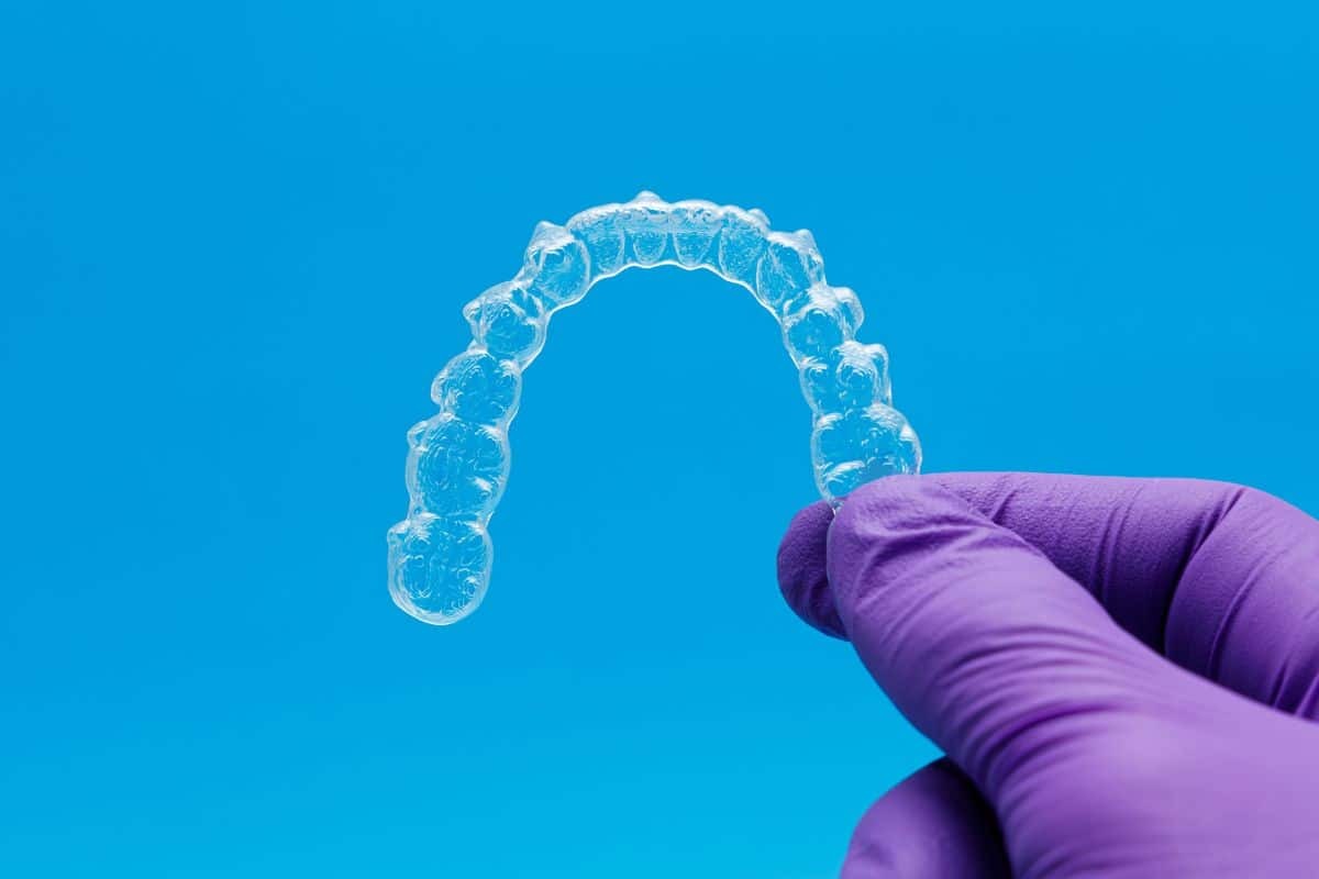 Clear dental aligner held in gloved hand against blue background, representing orthodontic treatment.