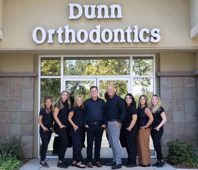 Nine members of the Dunn Orthodontics team, mostly in black attire, standing together outside the office entrance beneath the practice sign